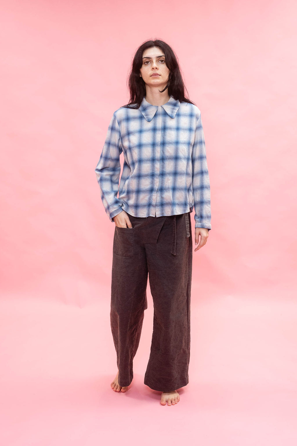 Person wearing a blue and white checkered shirt made from Japanese soft brushed cotton wool mix, and brown pants on a pink background
