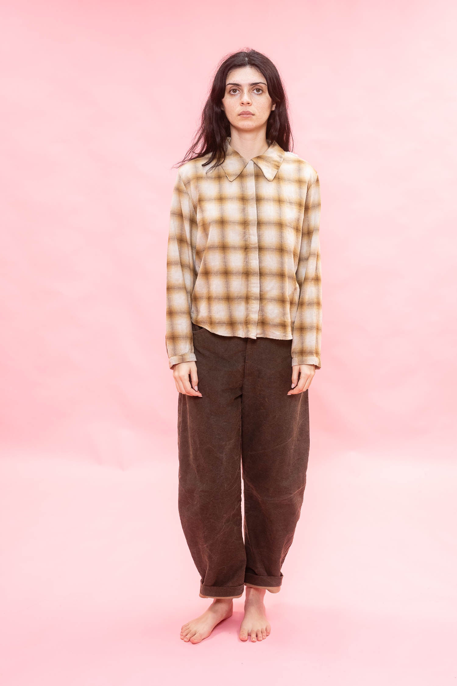 A woman stands barefoot against a solid pink studio background, wearing a long-sleeve button-up shirt with a tan and brown plaid pattern and loose dirt green cotton patina pants.