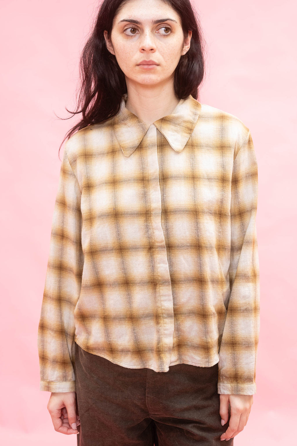 A woman wearing a cream and brown plaid button-down shirt with a pointed collar,  made with Japanese soft brushed cotton wool mix in ochre paired with dark brown pants, set against a solid pink background.
