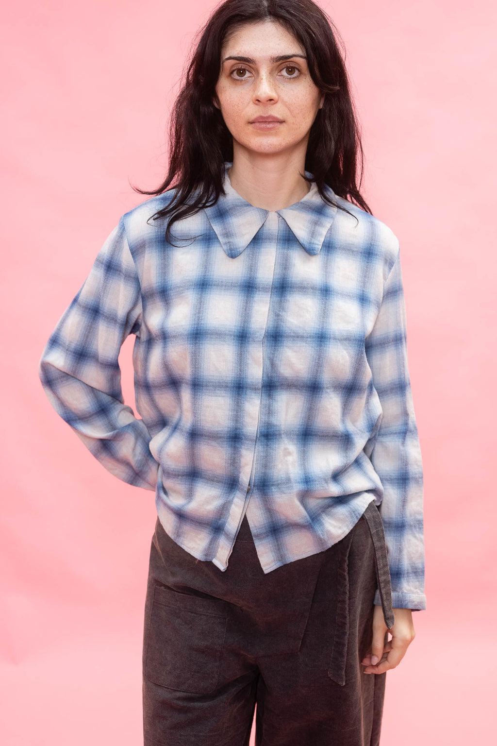 A woman modeling a long-sleeve button-up shirt with a blue and white plaid and a rounded collar, made with Japanese soft brushed cotton wool mix.  paired with dark brown pants, set against a solid pink background.
