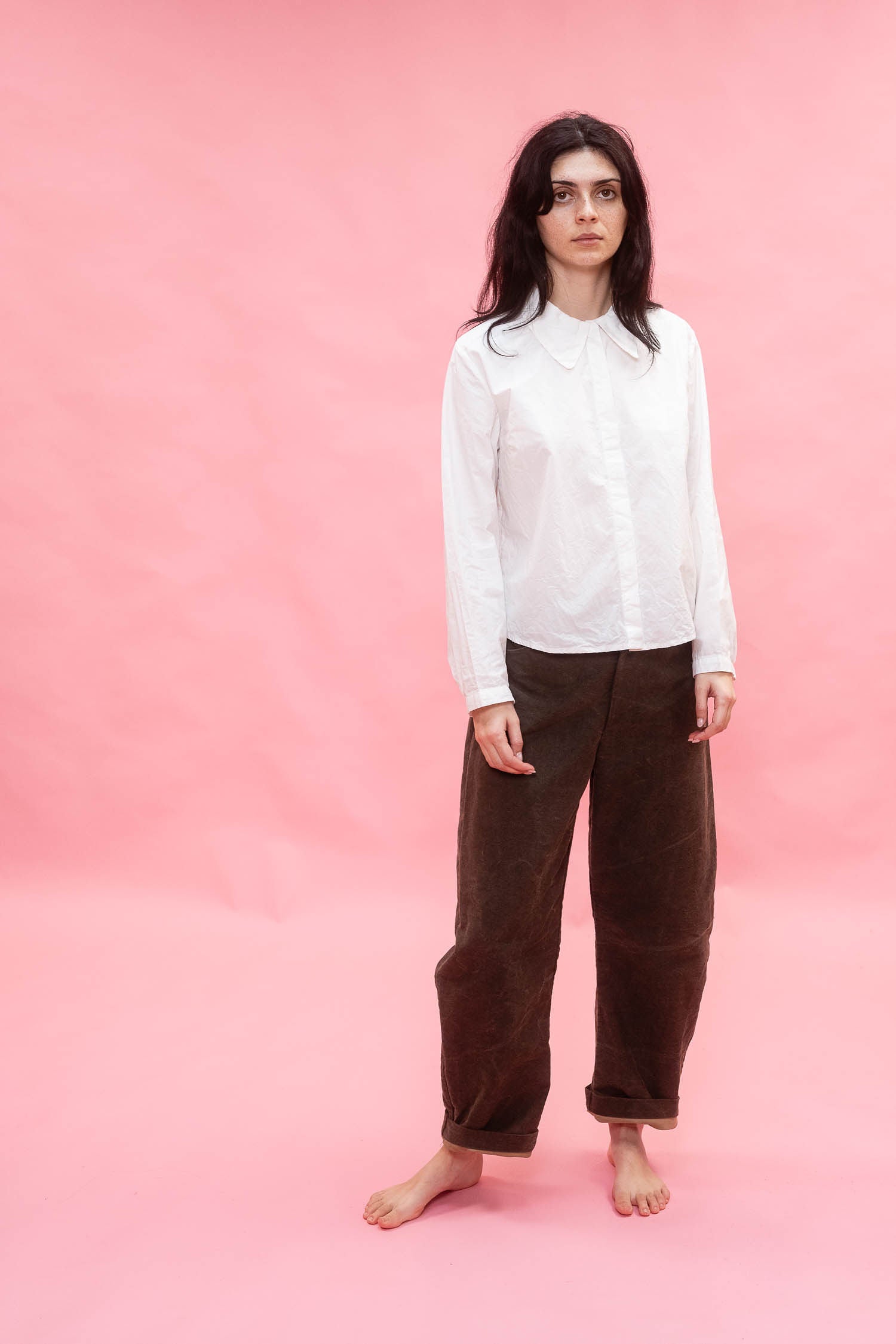 A woman stands barefoot against a solid pink studio backdrop, wearing a white long-sleeve button-up shirt and loose dirt green cotton patina pants.