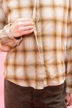 A close-up of a person wearing a long-sleeve button-up plaid shirt in beige, brown, and tan tones, made from Japanese soft brushed cotton wool mix, paired with dark brown pants, against a solid pink background.
