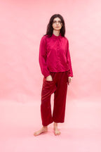 A woman stands barefoot against a solid pink studio background, wearing a shiny magenta long-sleeve button-up shirt and deep red wide-leg pants.