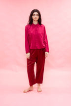 A woman stands barefoot against a solid pink studio backdrop, wearing a matching two-piece outfit consisting of a long-sleeve satin-like button-up shirt and relaxed-fit pants in a deep magenta-red color.