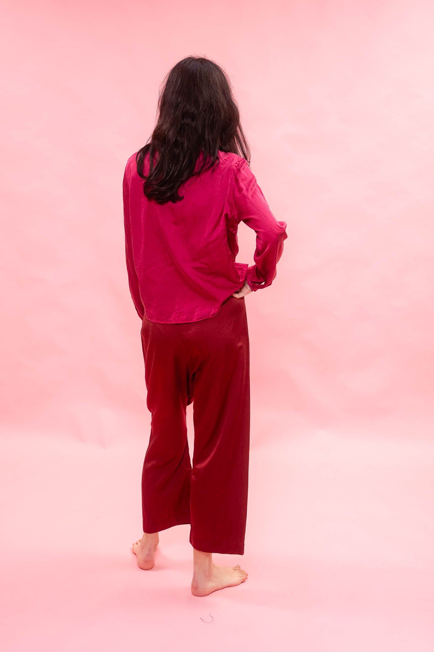 A woman stands with her back to the camera wearing a  lounge set in a vibrant magenta pink and deep red set against a solid light pink backdrop.