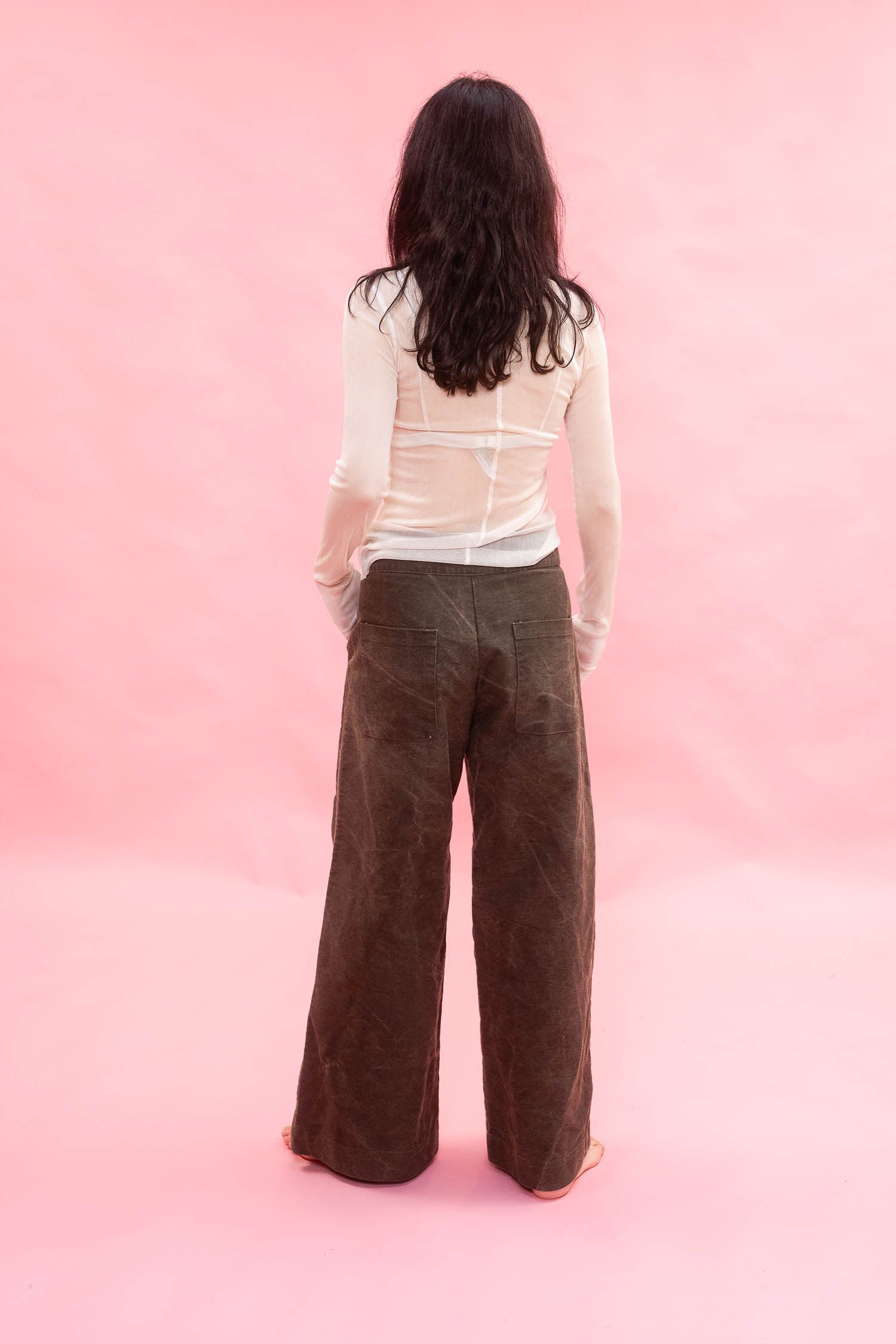 A woman viewed from behind wearing a white long-sleeve sheer top made from stretch knitted silk tulle, and dark brown wide-leg  pants, standing against a solid pink studio backdrop.
