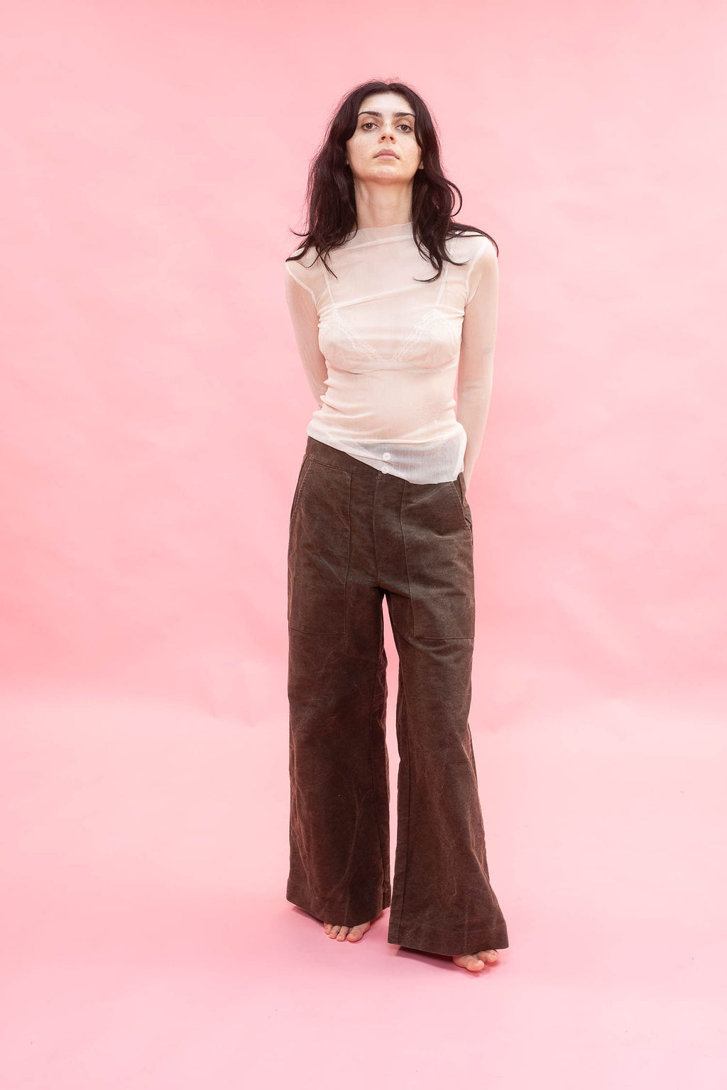 A woman stands against a solid pink studio backdrop wearing a sheer cream long-sleeve top made from stretch knitted silk tulle,and dark brown wide-leg corduroy pants.
