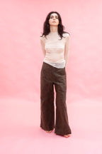 A woman stands against a solid pink studio backdrop wearing a sheer cream long-sleeve top made from stretch knitted silk tulle,and dark brown wide-leg corduroy pants.
