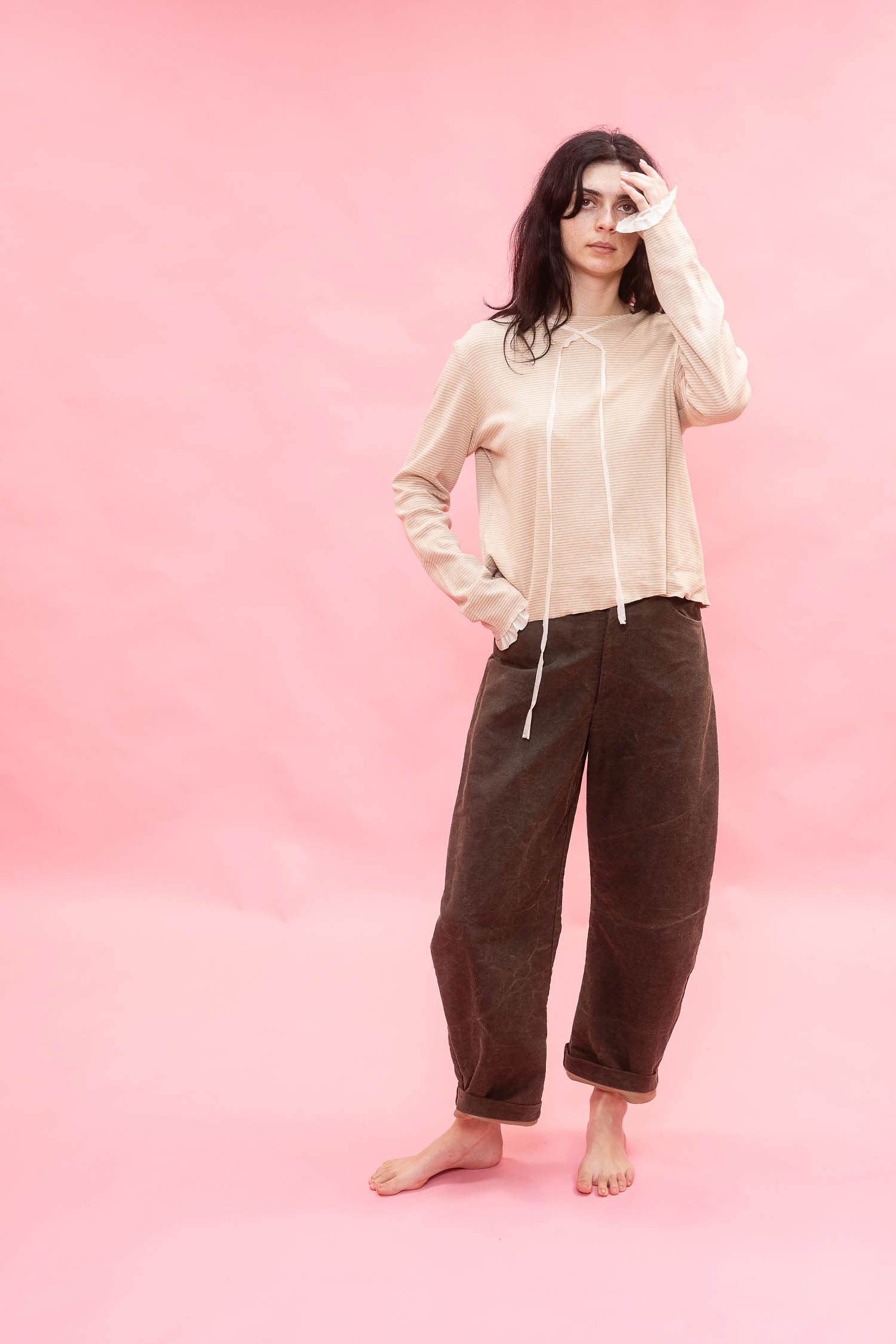 A woman stands barefoot against a solid pink backdrop, wearing a light beige long-sleeve striped t-shirt and dirt green cotton patina barrel legged pants.