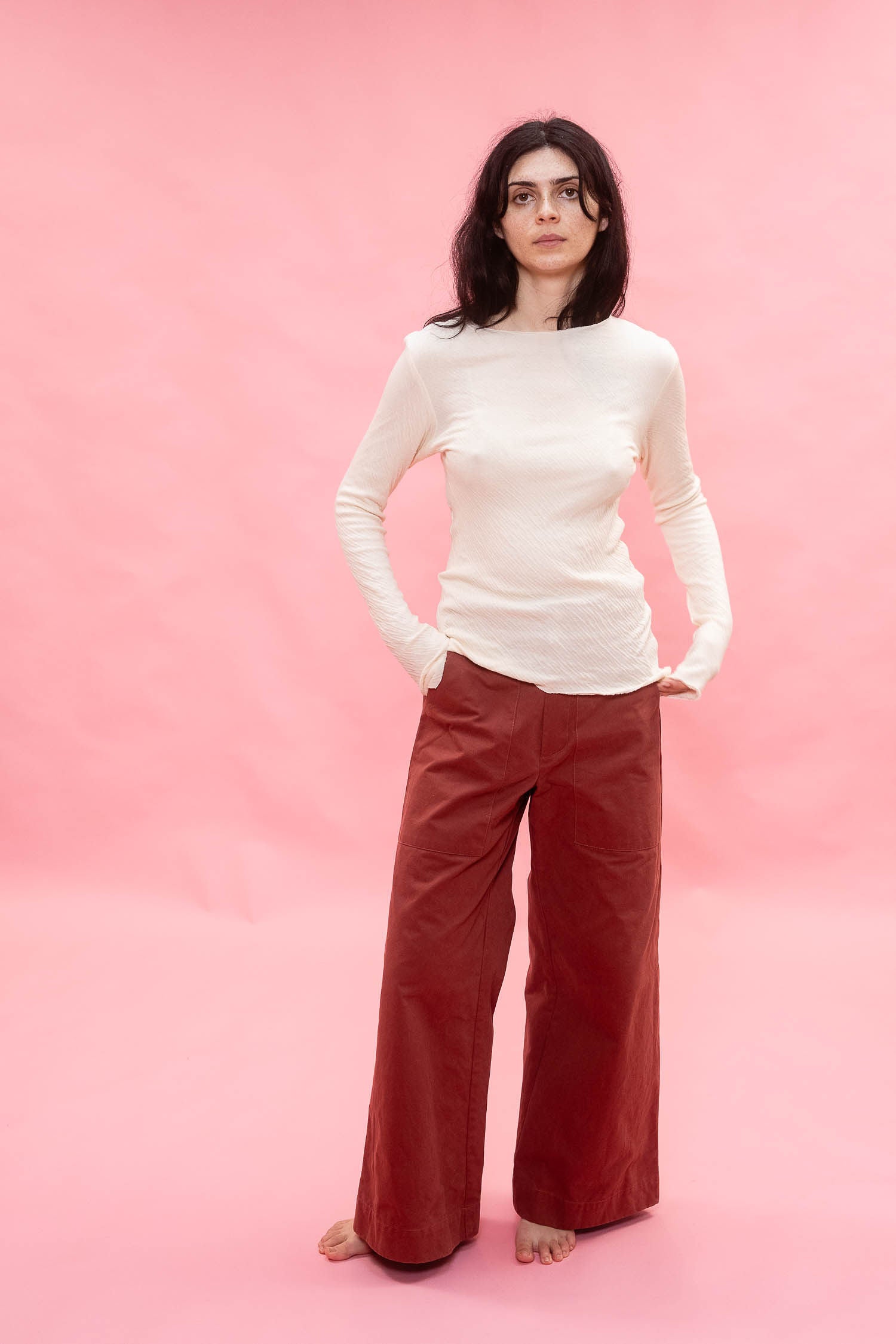A woman stands barefoot against a solid pink studio backdrop, wearing a fitted cream long-sleeve tee, cut from Japanese cotton double jersey, and rust red wide-leg pants.
