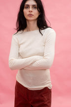 A woman with dark hair wearing a cream-colored, long-sleeve tee cut from Japanese cotton double jersey, and dark red pants, standing against a solid pink background with her arms crossed.
