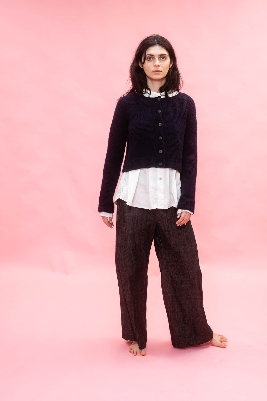 Person wearing a dark navy cashmere hand knit flower design cardigan, white shirt, and brown pants on a pink background
