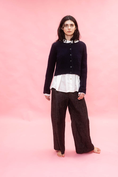 Person wearing a dark navy cashmere hand knit flower design cardigan, white shirt, and brown pants on a pink background
