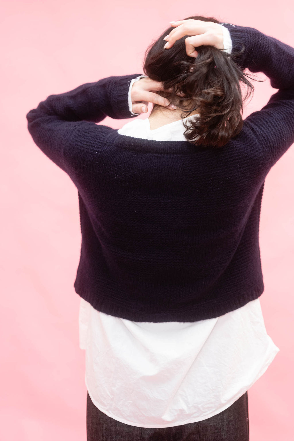 A woman shown from behind wearing a dark navy knit sweater over a white shirt, with their hands in their hair, set against a solid pink background.
