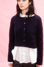 A woman wearing a dark navy hand knit cropped cashmere cardigan , with a flower design over a white collared shirt, set against a solid pink background.
