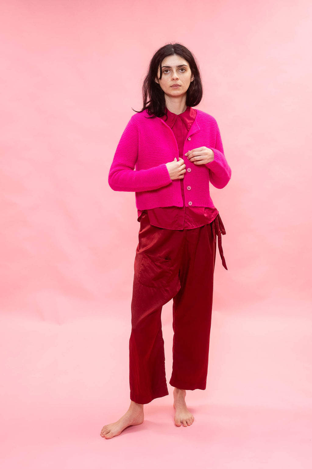 A model stands barefoot against a solid pink studio backdrop, wearing a bright fuchsia neon pink cashmere cropped hand knit button-front cardigan. the knit has a flower design, over silk top and deep burgundy wide-leg trousers.
