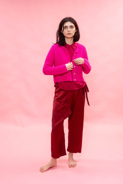 A model stands barefoot against a solid pink studio backdrop, wearing a bright fuchsia neon pink cashmere cropped hand knit button-front cardigan. the knit has a flower design, over silk top and deep burgundy wide-leg trousers.
