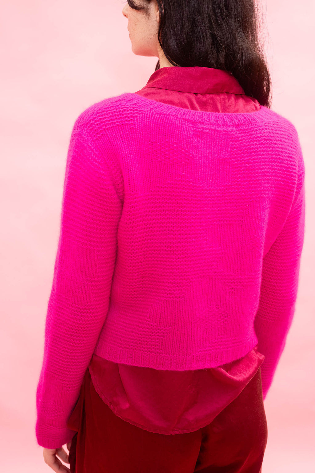 A woman shown from the back wearing a bright hot neon pink hand knitted cashmere with a flower design, cropped cardigan over a darker pink top, set against a solid light pink background.
