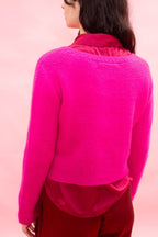 A woman shown from the back wearing a bright hot neon pink hand knitted cashmere with a flower design, cropped cardigan over a darker pink top, set against a solid light pink background.
