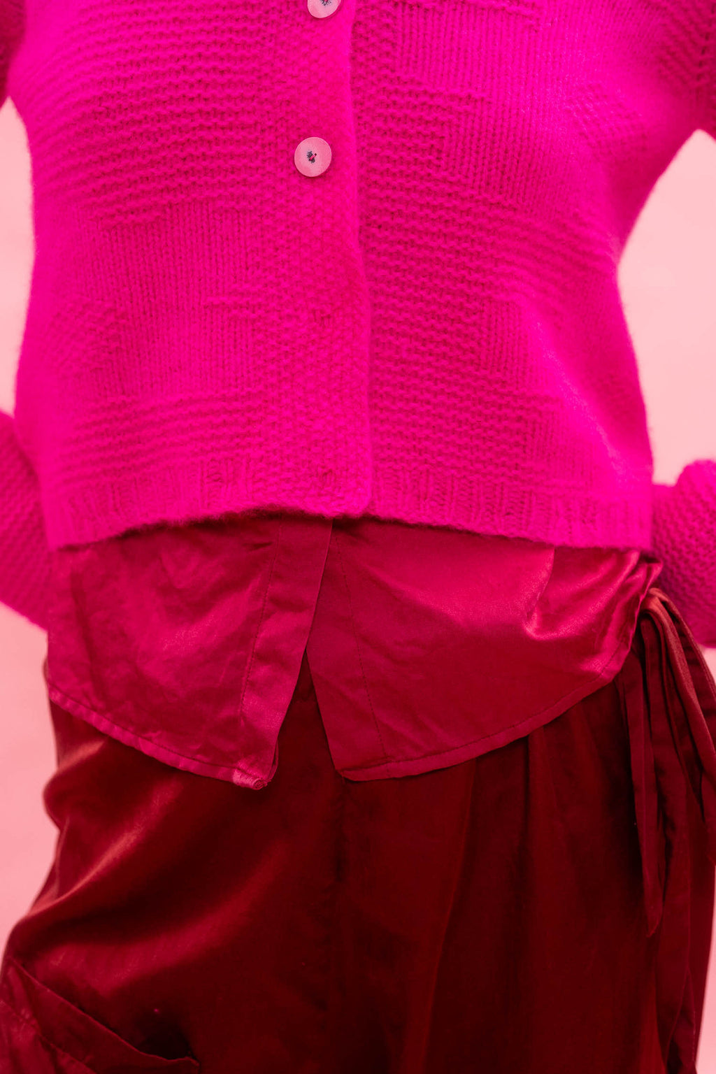 Person wearing a bright neon pink hand knitted cashmere cardigan over a red outfit on a pink background
