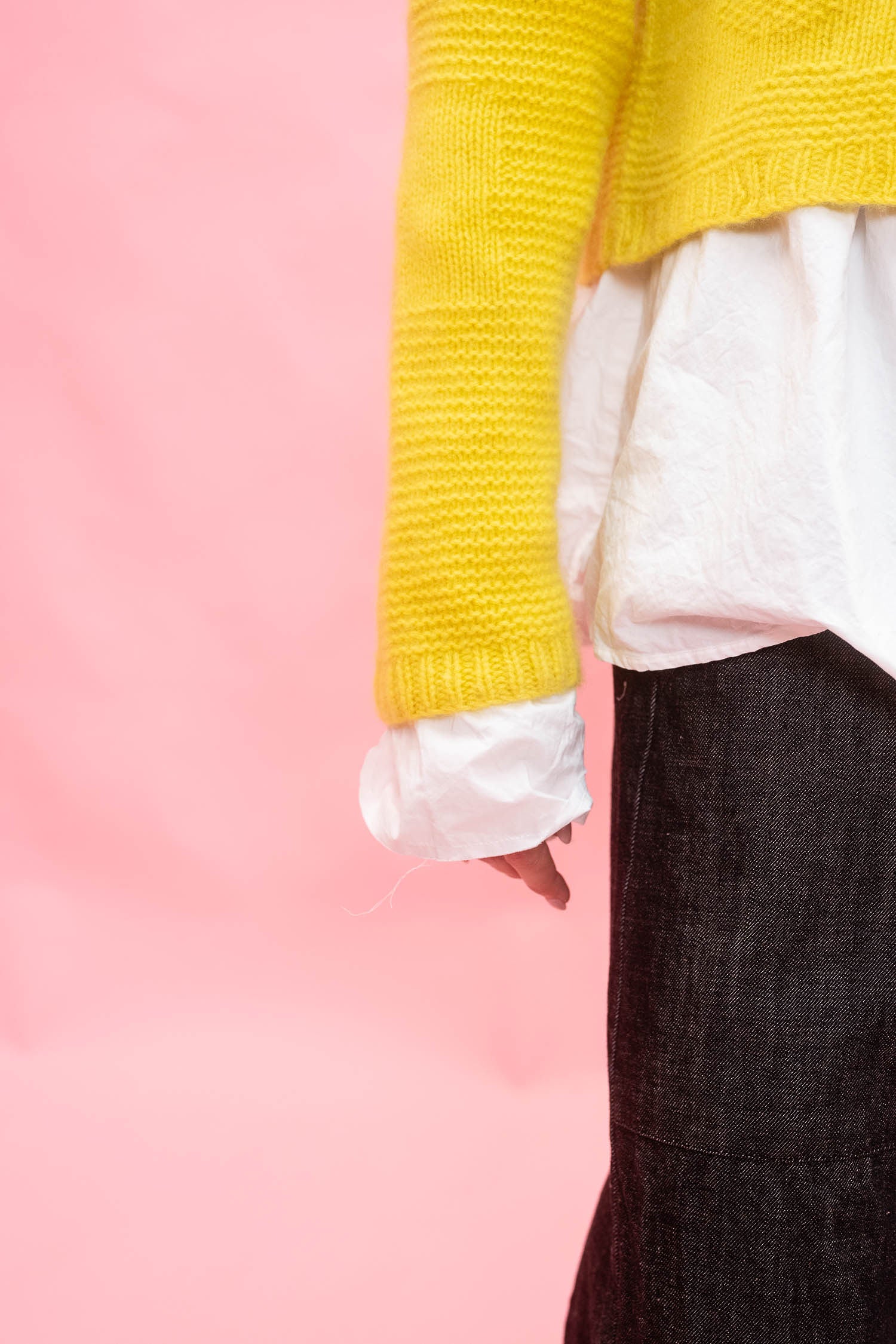 A close-up of a person wearing a bright yellow flower design hand knit cashmere cardigan over a white top and dark gray skirt, set against a solid pink background.
