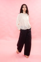 Woman wearing a sheer cream Organdy blouse and black pants on a pink background