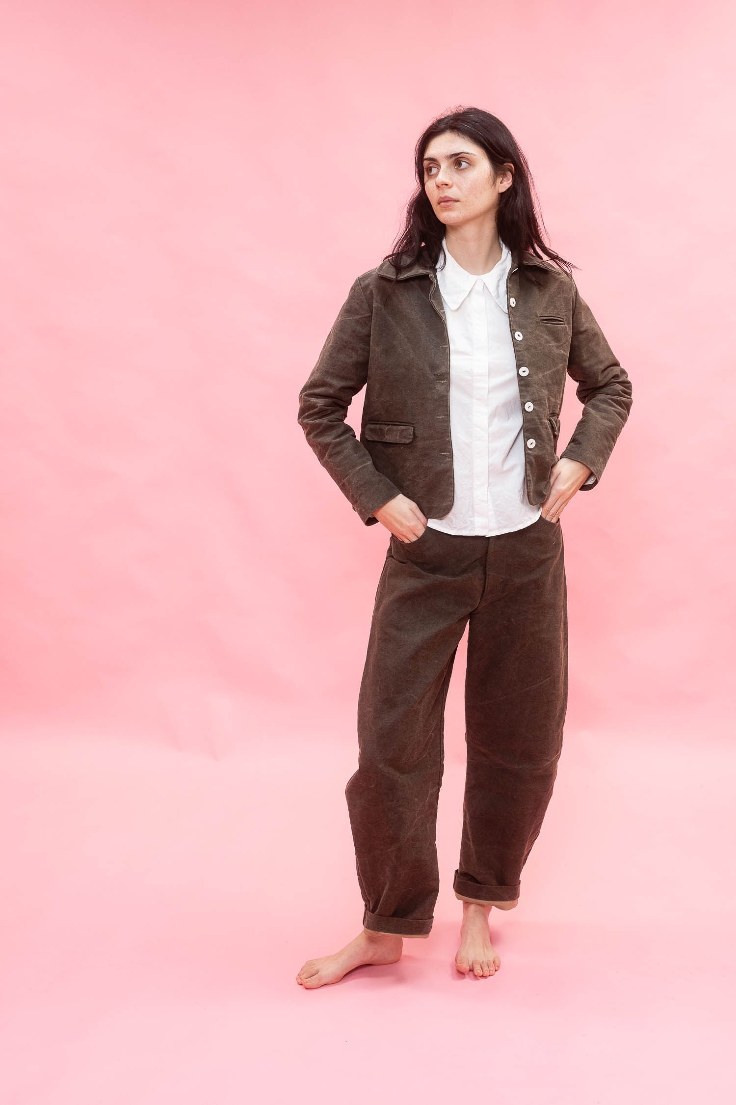 A woman stands barefoot against a solid pink studio background, wearing a dirt green cotton patina jacket and matching trousers over a white collared shirt.