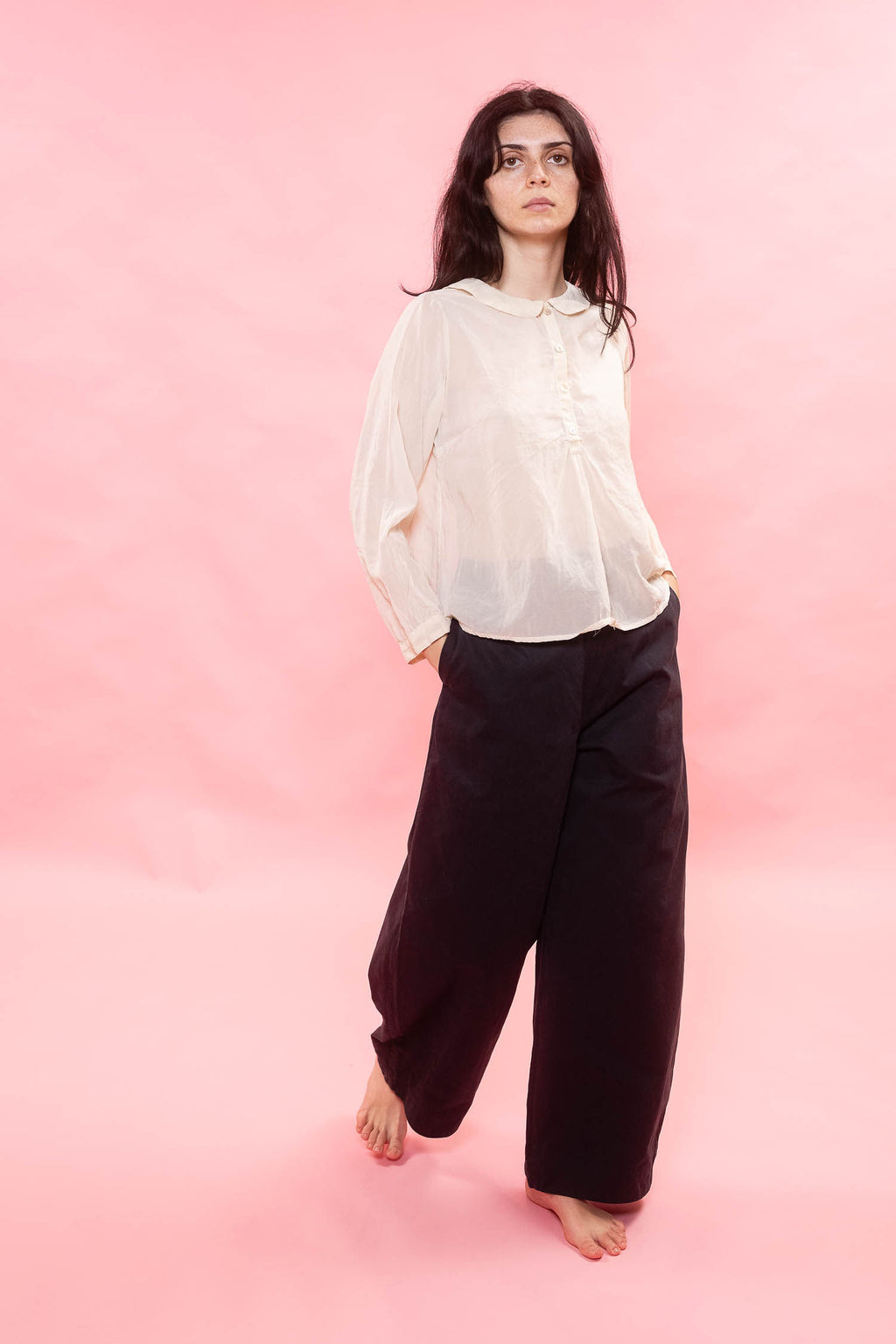 Woman wearing a sheer cream organdy blouse and black pants on a pink background
