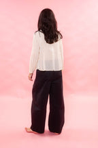 A woman shown from behind wearing a cream organdie long-sleeve sheer blouse and loose black wide-leg pants, standing against a solid pink studio background.
