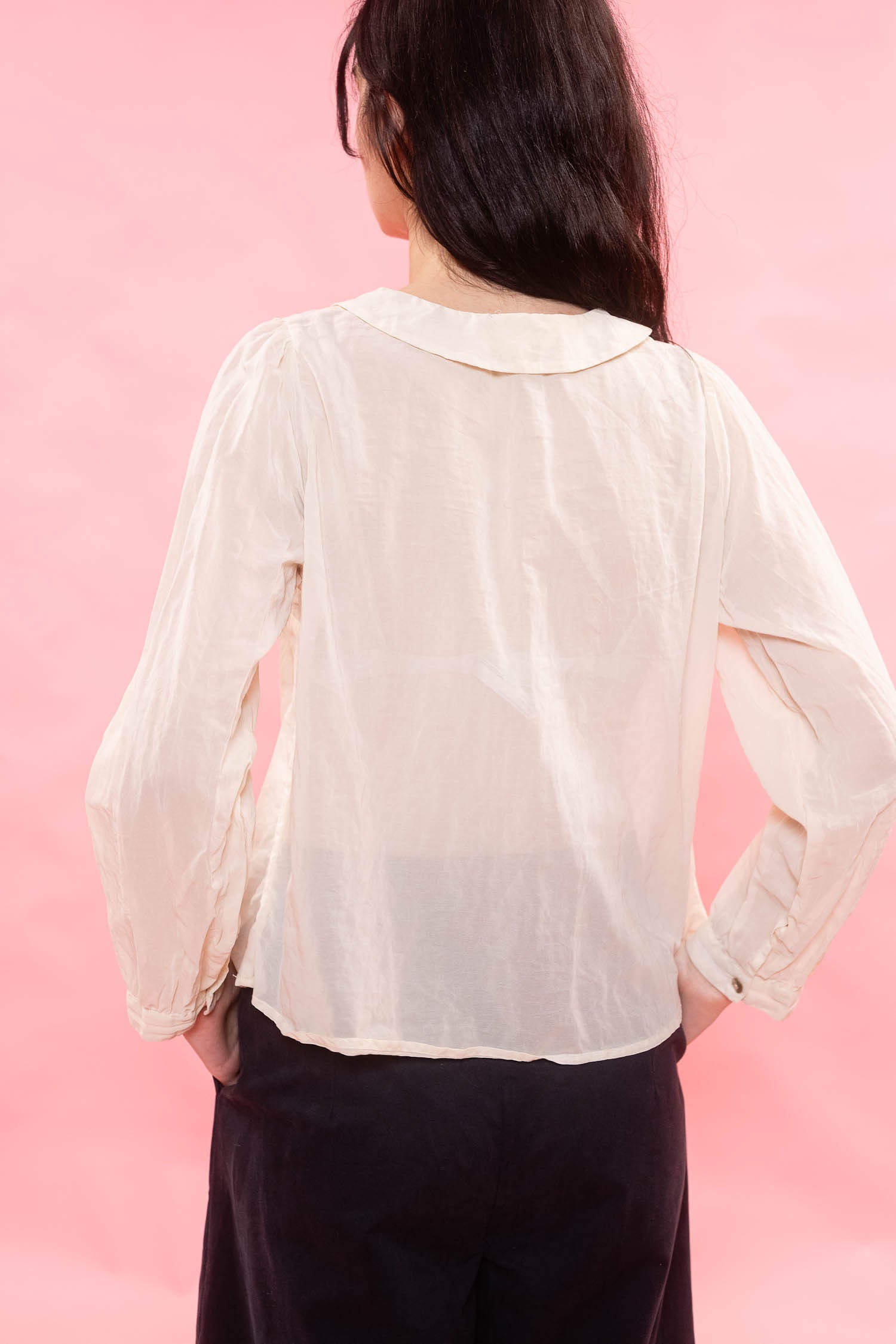 A woman viewed from the back wearing a sheer, long-sleeve cream blouse and dark pants, set against a solid pink background.
