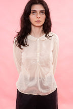 A woman with dark hair wearing a semi-sheer, cream organdy long-sleeve blouse with a Peter Pan collar and front buttons, paired with dark trousers, set against a solid pink background.
